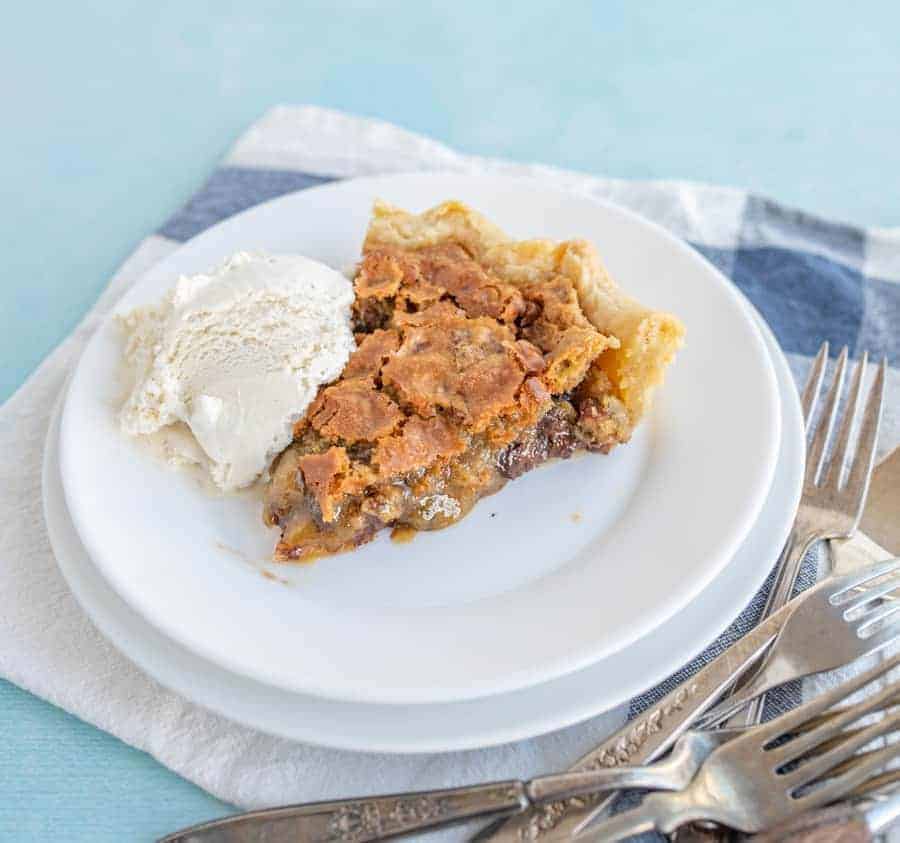 A delicious looking piece of pie is on a white pie plate snuggled up to a scoop of vanilla ice cream. There are forks sticking around the bottom right side of the plate on top of a blue and white checkered towel.