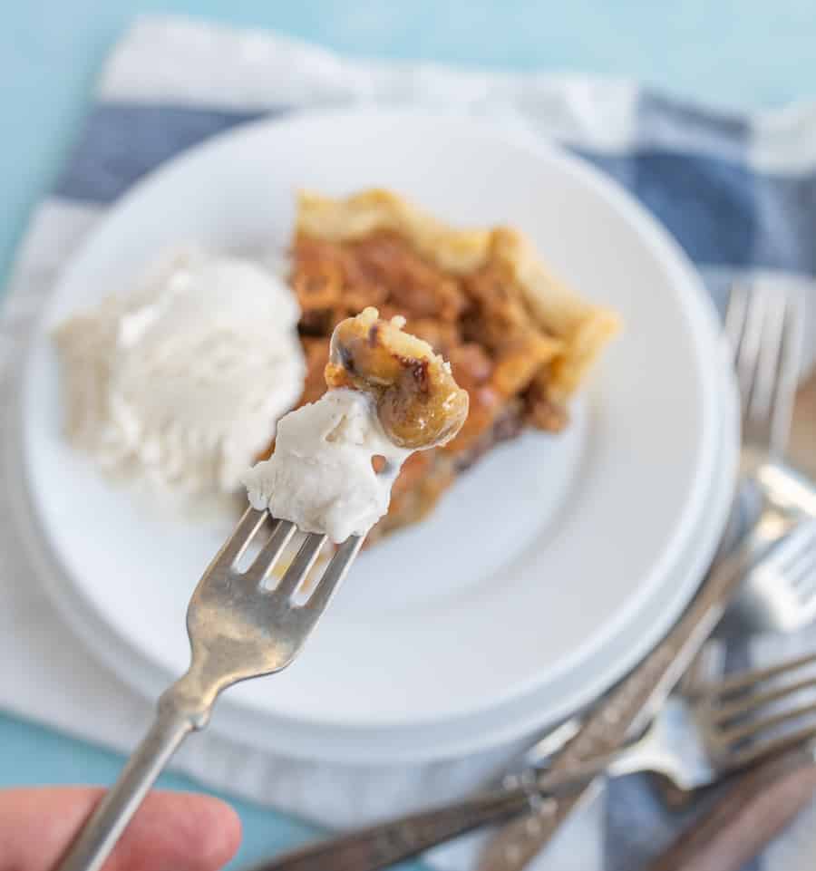 In the foreground, there is a piece of pie and a bite of vanilla ice cream on a fork being held with a finger showing. In the background is a piece of pie and a scoop of vanilla ice cream on a plate on top of a blue and white checkered towel and forks around the bottom right side.