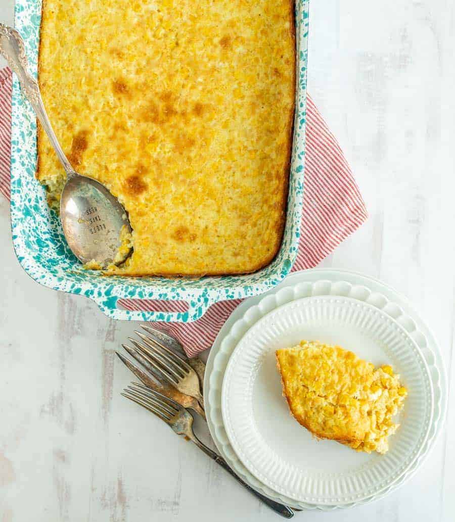 An overhead shot with a pan full of corn pudding, except a scoop taken out in the bottom left-hand side. In that spot is a silver spoon embossed with "Bless this Mess." In the bottom right corner is a serving of the corn pudding in the middle of a small white plate that is stacked on top of a couple more white plates with varying designs. to the left of the plates are some silver forks at a 45 degree angle.