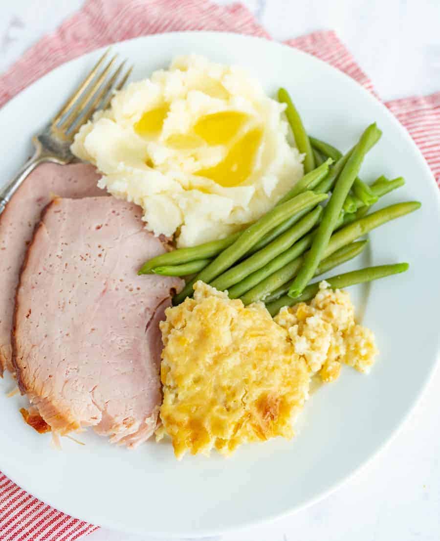 An up-close shot of a white dinner plate with corn pudding in the bottom right, green beans above that, with mashed potatoes complete with butter melting on them in the top middle. On the bottom left are two slices of ham, and a silver fork by them. The white plate is sitting on top of a towel with skinny red and white stripes.