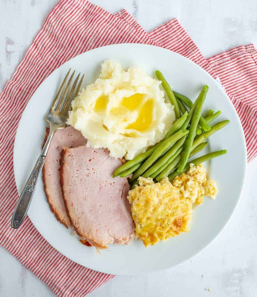An above shot that shows an entire white dinner plate, complete with corn pudding in the bottom right, green beans above that, mashed potatoes with butter melting on them in the upper middle, ham in the bottom left, and a fork next to the ham and potatoes. Underneath the white plate is a skinny striped red and white towel.