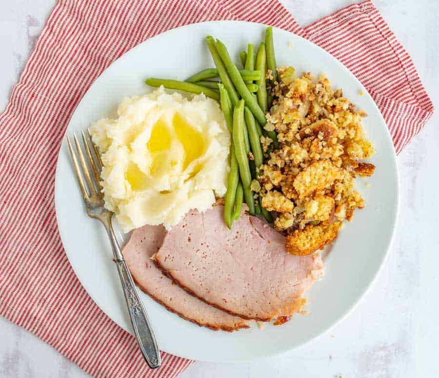 A white dinner plate has a silver fork angled from the middle of the bottom toward the left upper corner with mashed potatoes that have butter melting on them, green beans, stuffing, and two slices of ham. The plate is sitting on a skinny striped red and white dish towel.