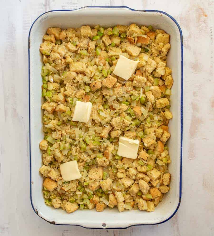 A white rectangular pan with blue outline is filled with pieces of dried out bread, celery, and other spices and four slices of butter rest on top.