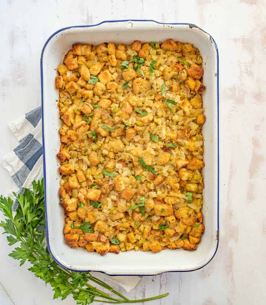 Cooked stuffing with browned bread crumbs, celery and other spices are in a rectangular white dish with a blue outline resting on a blue and white checkered towel and garnished by some parsley.