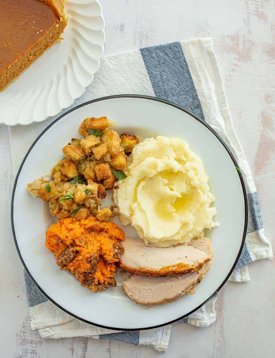 A white dinner plate with a dark green outline is filled with stuffing, mashed potatoes with butter melting on top, a few slices of roasted turkey, and some sweet potato casserole are resting on a white dish towel with blue stripes and part of a piece of pumpkin pie is resting on a fancy white salad plate.