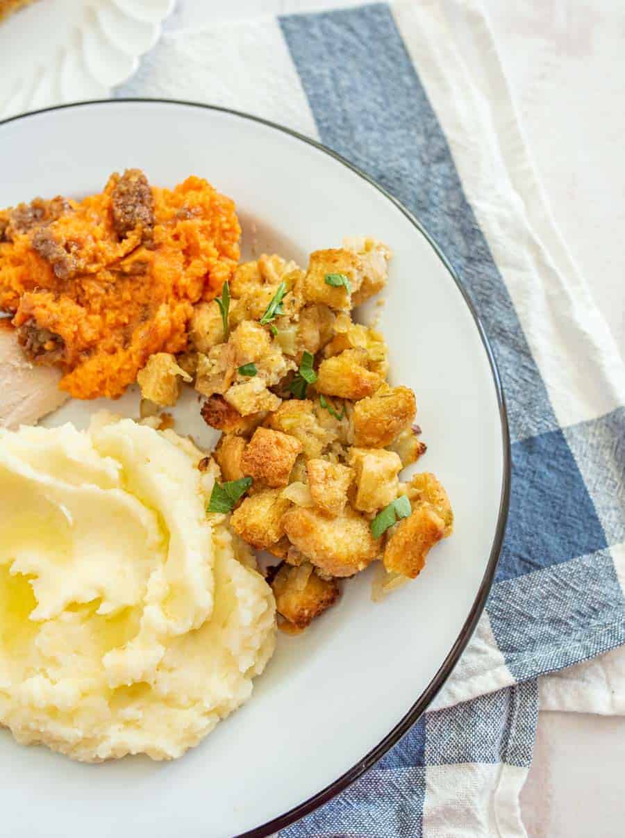 Most of a white dinner plate with a dark green outline is shown. The dinner plate has mashed potatoes with butter melting on them, a small piece of turkey can be seen, a serving of sweet potato casserole, and stuffing are also on the plate. Underneath the plate is a white dishtowel with a blue stripe and in the upper left hand corner, you can see a slice of pumpkin pie on a fancy white salad plate.