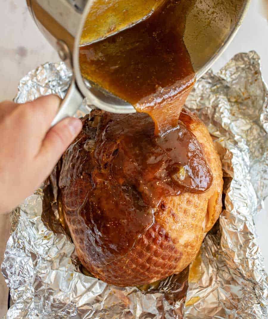 The perfectly cooked, browned ham is sitting in the tinfoil that had covered it while being cooked and a hand is holding a sauce pan full of glaze that is cascading out of the pan and down the ham.