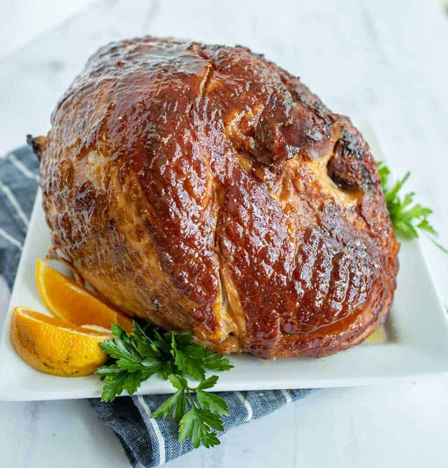 A perfectly cooked ham, covered in glaze is garnished by some parsley and orange slices on a white square dish on top of a blue with small white striped hand towel.