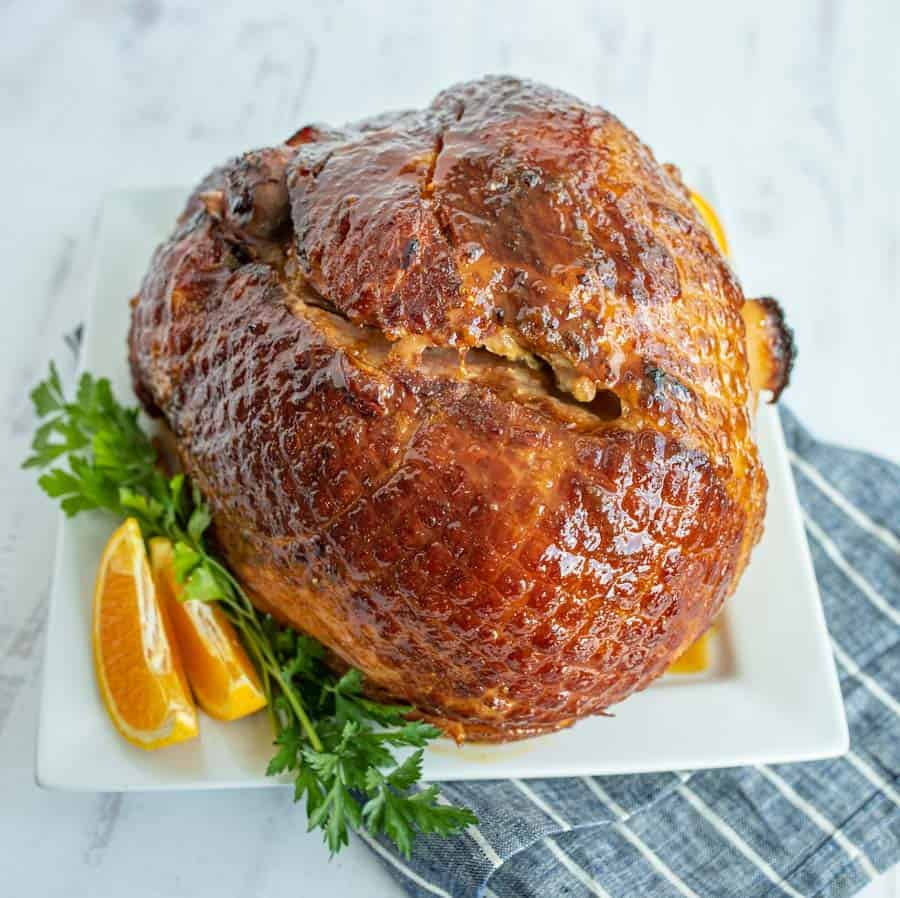 Another angle of the perfectly cooked ham, this picture is taken from a little bit higher up. The ham is browned with glaze and garnished by two orange slices and parsley on a square white dish on top of a blue towel that has thin white stripes on it.