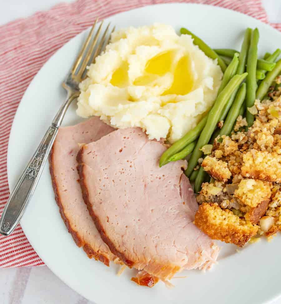 A white plate has a silver fork angled on it next to mashed potatoes that have butter melting on them, green beans, stuffing, and two slices of ham. The plate is on top of a skinny striped red and white towel.