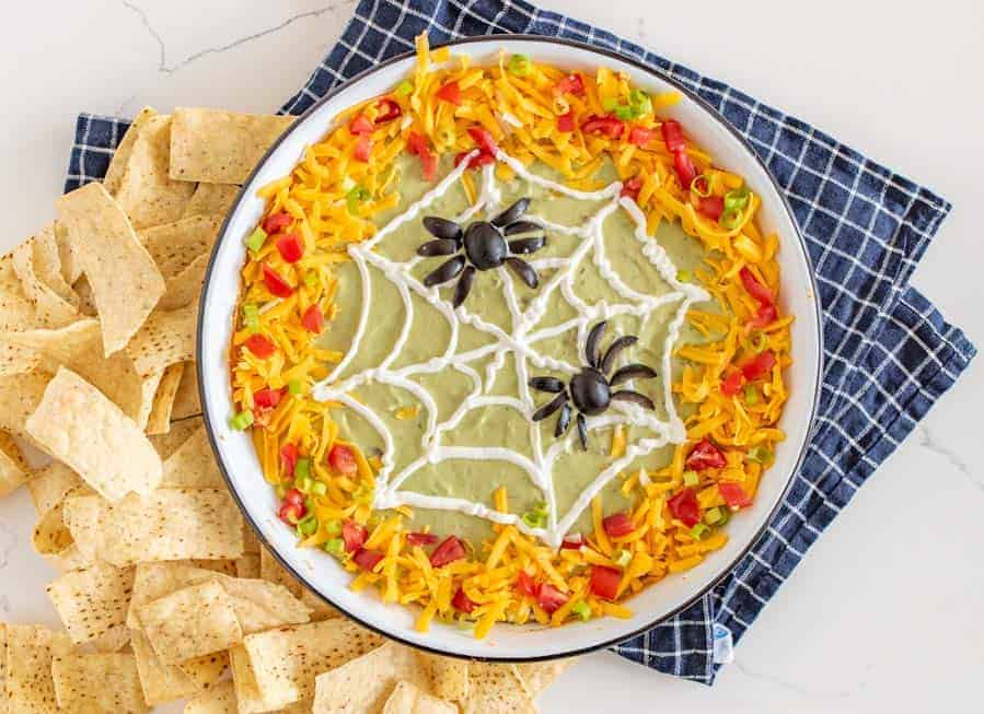 This easy Halloween bean dip is a no-bake recipe full of delicious ingredients that is SO easy to make, and its simple sour cream spider web makes it perfect to bring along to all your Halloween festivities! Wondering what to bring to this year's Halloween party? This bean dip is such a breeze to whip up (no cooking involved) and it looks intricate even though it's so easy, fast, and tasty! #beandip #layeredbeandip #refriedbeans #halloweenrecipes #halloweenbeandip #chipsanddip