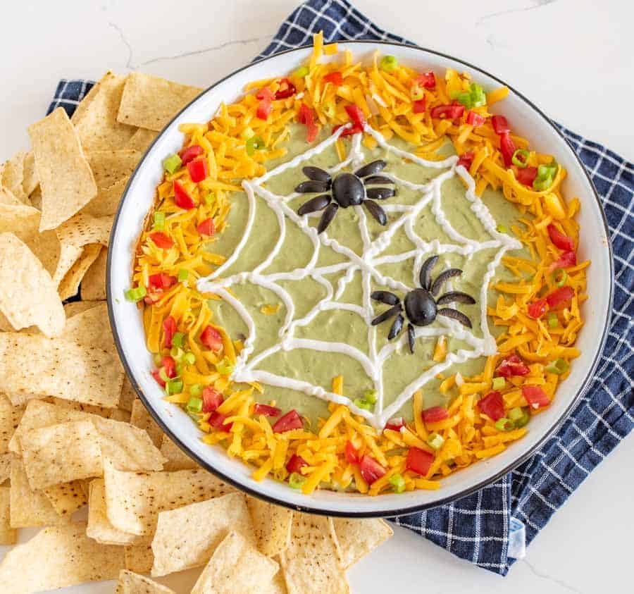 This easy Halloween bean dip is a no-bake recipe full of delicious ingredients that is SO easy to make, and its simple sour cream spider web makes it perfect to bring along to all your Halloween festivities! Wondering what to bring to this year's Halloween party? This bean dip is such a breeze to whip up (no cooking involved) and it looks intricate even though it's so easy, fast, and tasty! #beandip #layeredbeandip #refriedbeans #halloweenrecipes #halloweenbeandip #chipsanddip