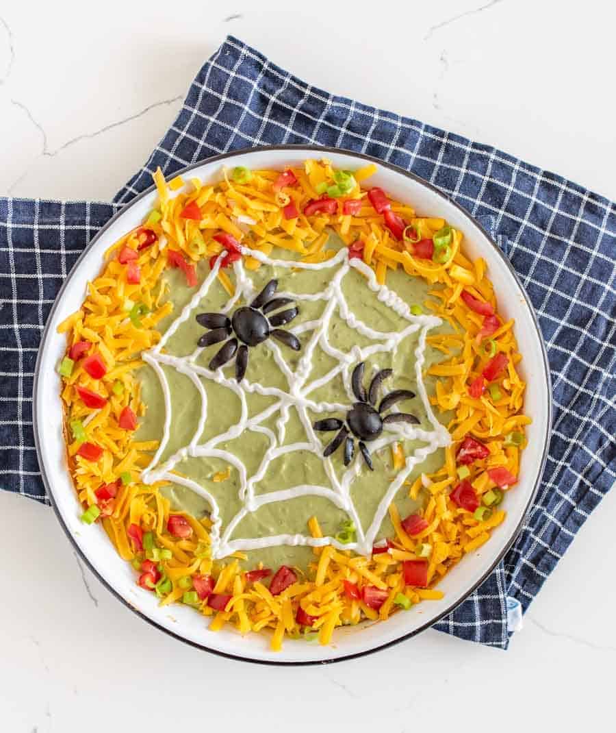 This easy Halloween bean dip is a no-bake recipe full of delicious ingredients that is SO easy to make, and its simple sour cream spider web makes it perfect to bring along to all your Halloween festivities! Wondering what to bring to this year's Halloween party? This bean dip is such a breeze to whip up (no cooking involved) and it looks intricate even though it's so easy, fast, and tasty! #beandip #layeredbeandip #refriedbeans #halloweenrecipes #halloweenbeandip #chipsanddip