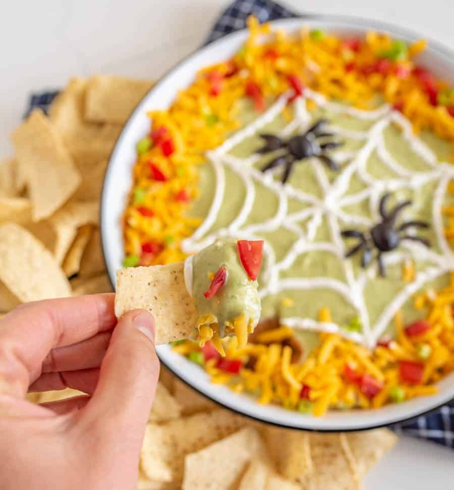 This easy Halloween bean dip is a no-bake recipe full of delicious ingredients that is SO easy to make, and its simple sour cream spider web makes it perfect to bring along to all your Halloween festivities! Wondering what to bring to this year's Halloween party? This bean dip is such a breeze to whip up (no cooking involved) and it looks intricate even though it's so easy, fast, and tasty! #beandip #layeredbeandip #refriedbeans #halloweenrecipes #halloweenbeandip #chipsanddip