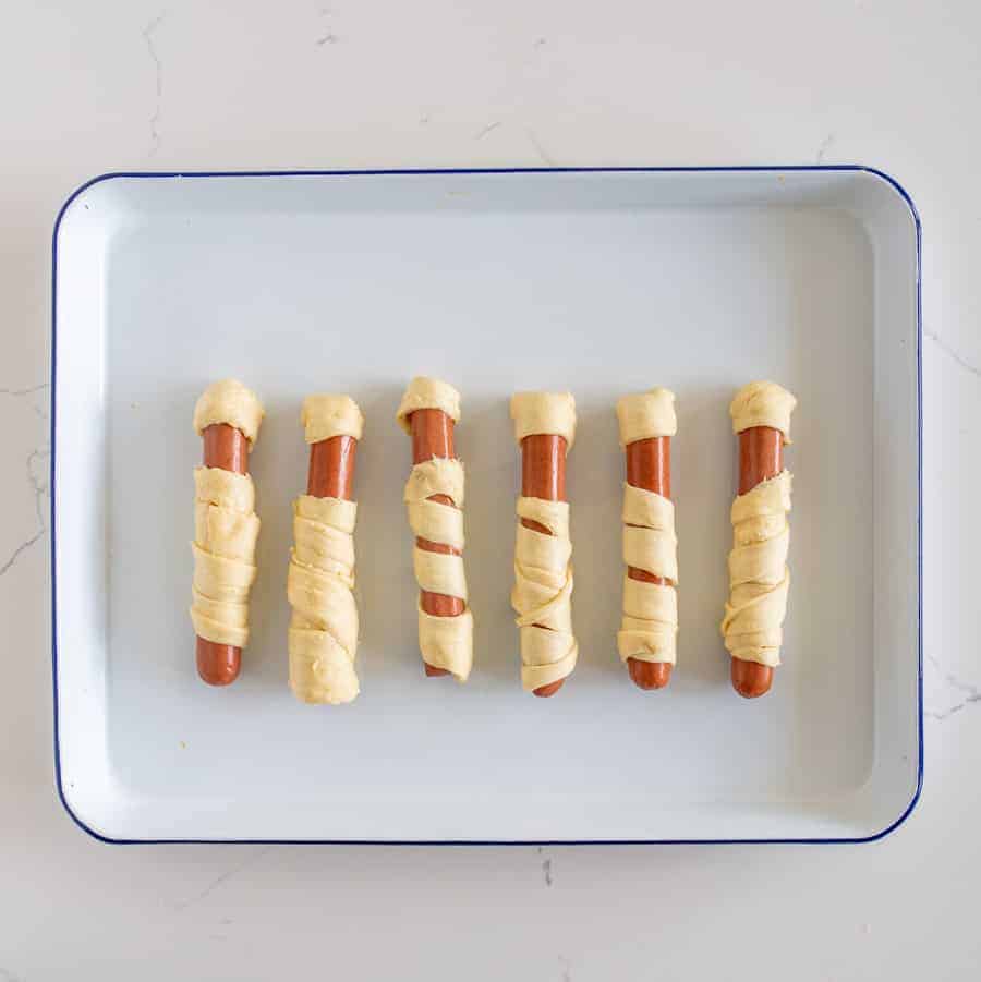 a white tray with a blue rim and six crescent dough-wrapped hot dogs.