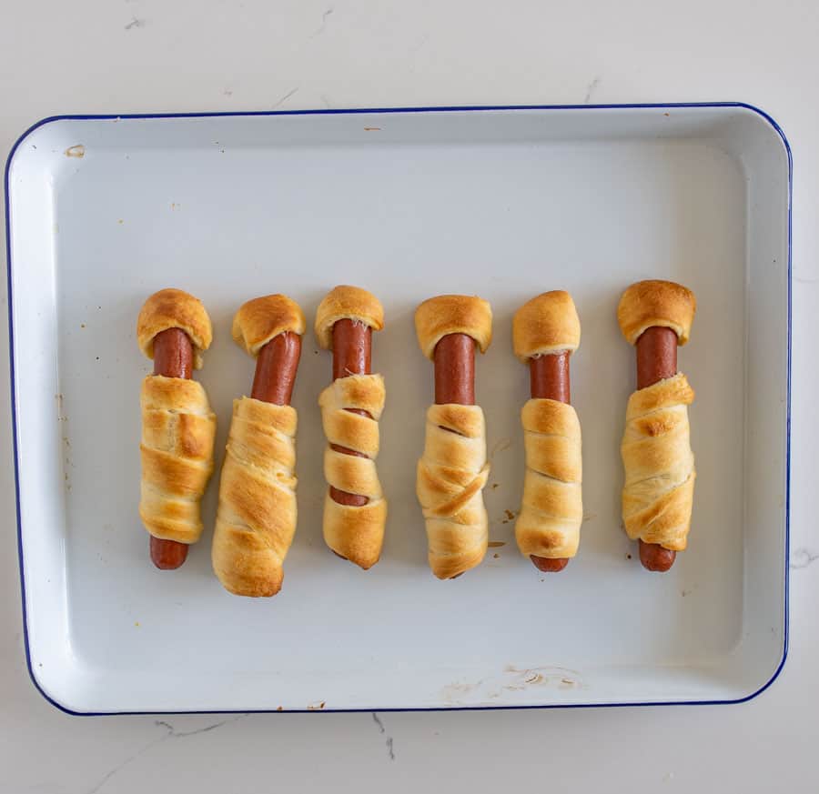 a white tray with a blue rim and six crescent dough-wrapped hot dogs that have been baked.