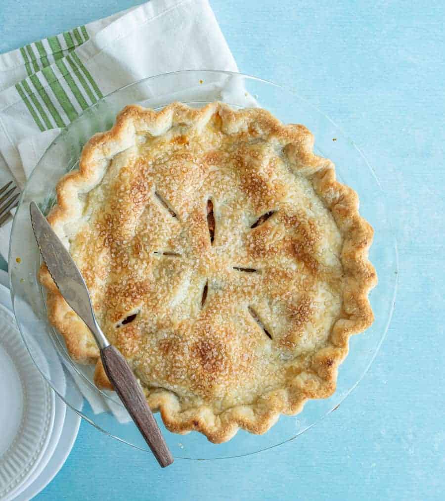 A golden-brown, homemade pie with a flaky crust sits in a glass pie dish on a light blue surface. A knife and pie server rest on top, with plates and a white napkin nearby.