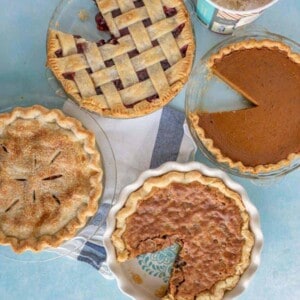 Four different pies on a blue surface, including a lattice-topped cherry pie, a pumpkin pie, an apple pie with a full top crust, and a pecan pie with a slice missing. A tub of ice cream is nearby.