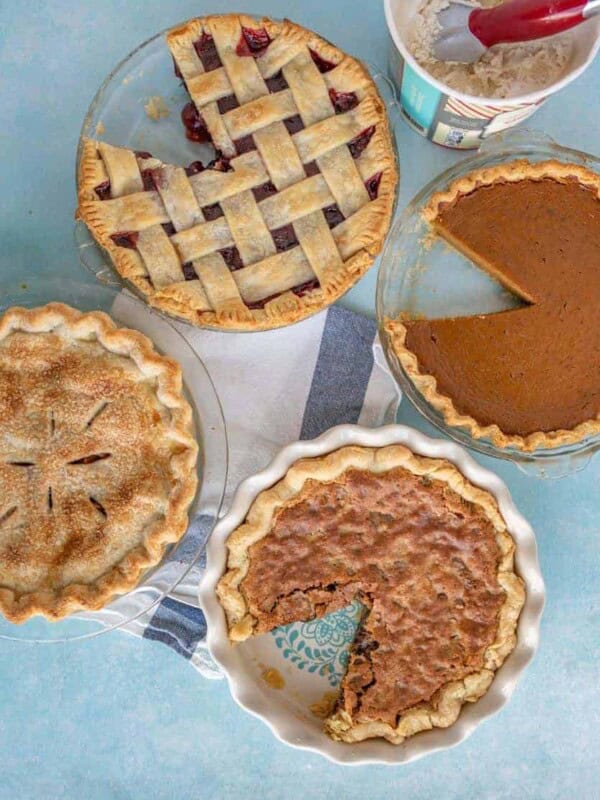 Four different pies on a blue surface, including a lattice-topped cherry pie, a pumpkin pie, an apple pie with a full top crust, and a pecan pie with a slice missing. A tub of ice cream is nearby.