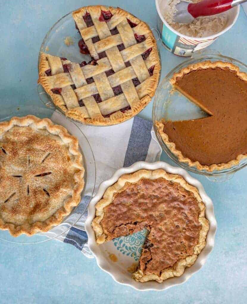 Four different pies on a blue surface, including a lattice-topped cherry pie, a pumpkin pie, an apple pie with a full top crust, and a pecan pie with a slice missing. A tub of ice cream is nearby.