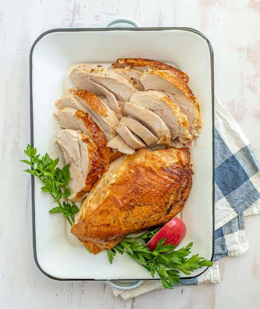 The roasted turkey breast is now cut up with parsley and a red apple slice to garnish it inside of a white pan on top of a blue and white checkered dish towel.
