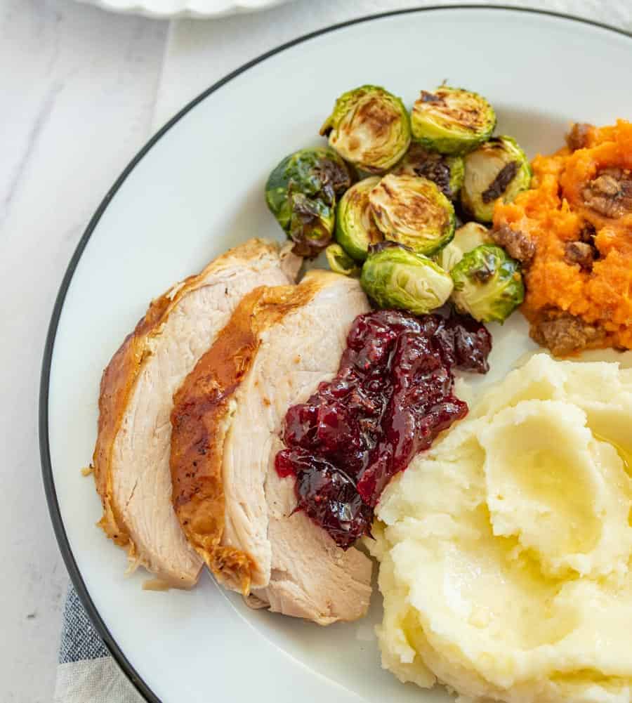 Two slices of cooked turkey breast with some cranberry sauce on it next to mashed potatoes, yams, and artichokes on a white dinner plate outlined with green.