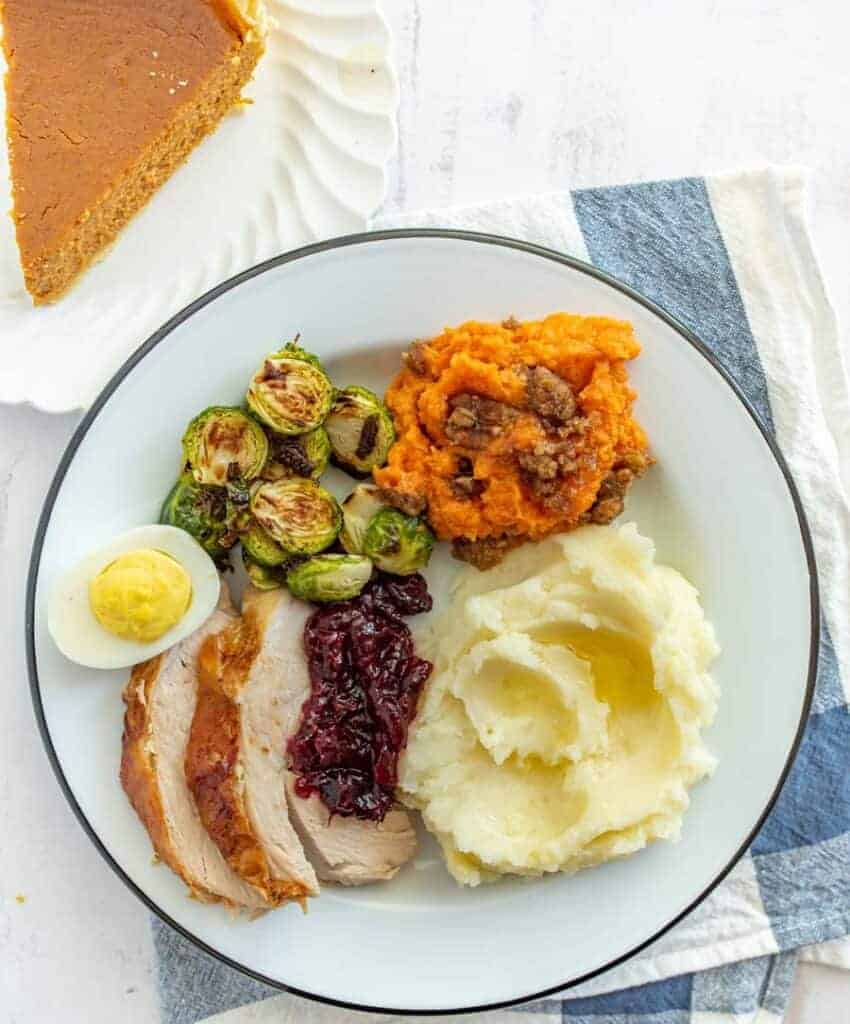 A dinner plate features turkey slices with cranberry sauce, mashed potatoes with gravy, roasted Brussels sprouts, a streusel-topped sweet potato casserole with pecans, and half a deviled egg. A slice of pumpkin pie is on a separate plate.