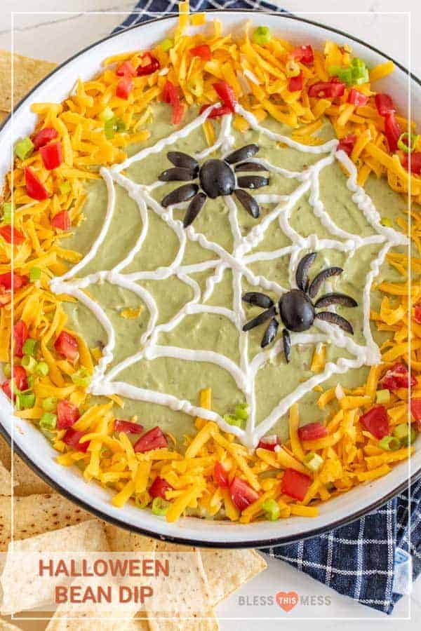 A bowl of Halloween Bean Dip with a Spider Web and Spider decoration atop guacamole, shredded cheese, diced tomatoes and scallions