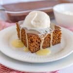 A square piece of gingerbread cake on a white plate topped with vanilla caramel sauce and whipped cream