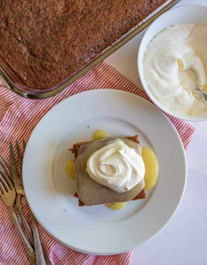 A square piece of gingerbread cake on a white plate topped with vanilla caramel sauce and whipped cream