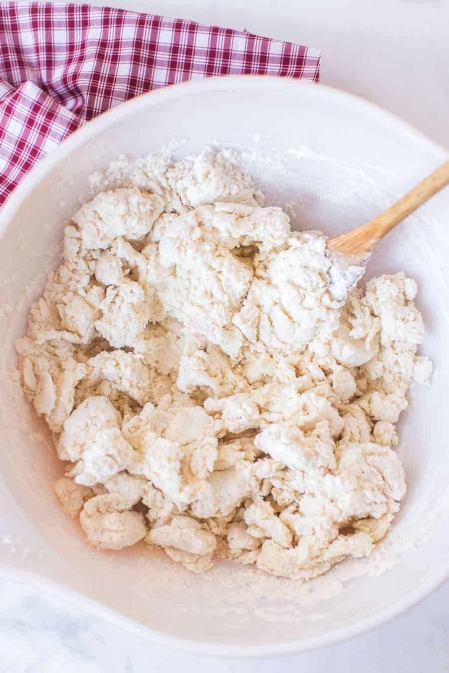 simple cream biscuit dough in white mixing bowl with wooden spoon on red and white checked towel