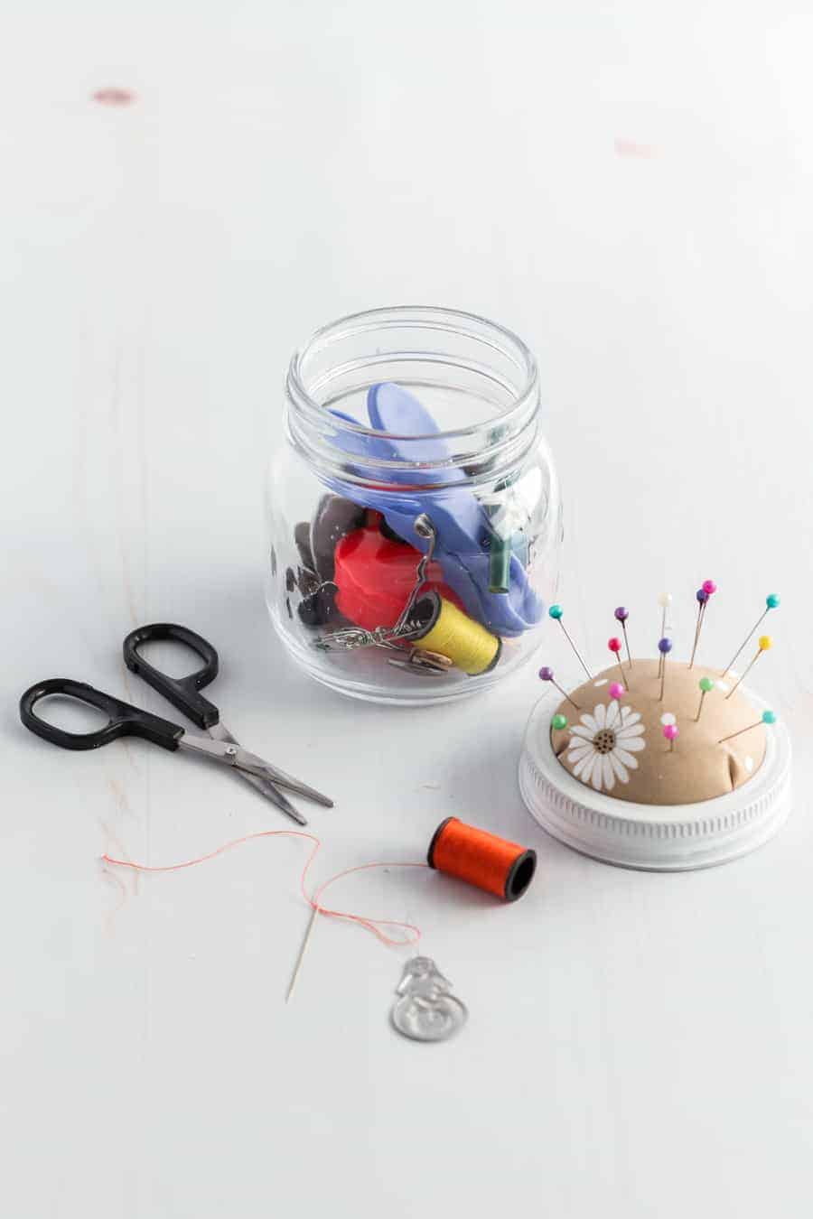 A jar full of sewing tools with scissors out by it, a threaded needle connected to the spool, a threader, and the jar lid made into a pin cushion.