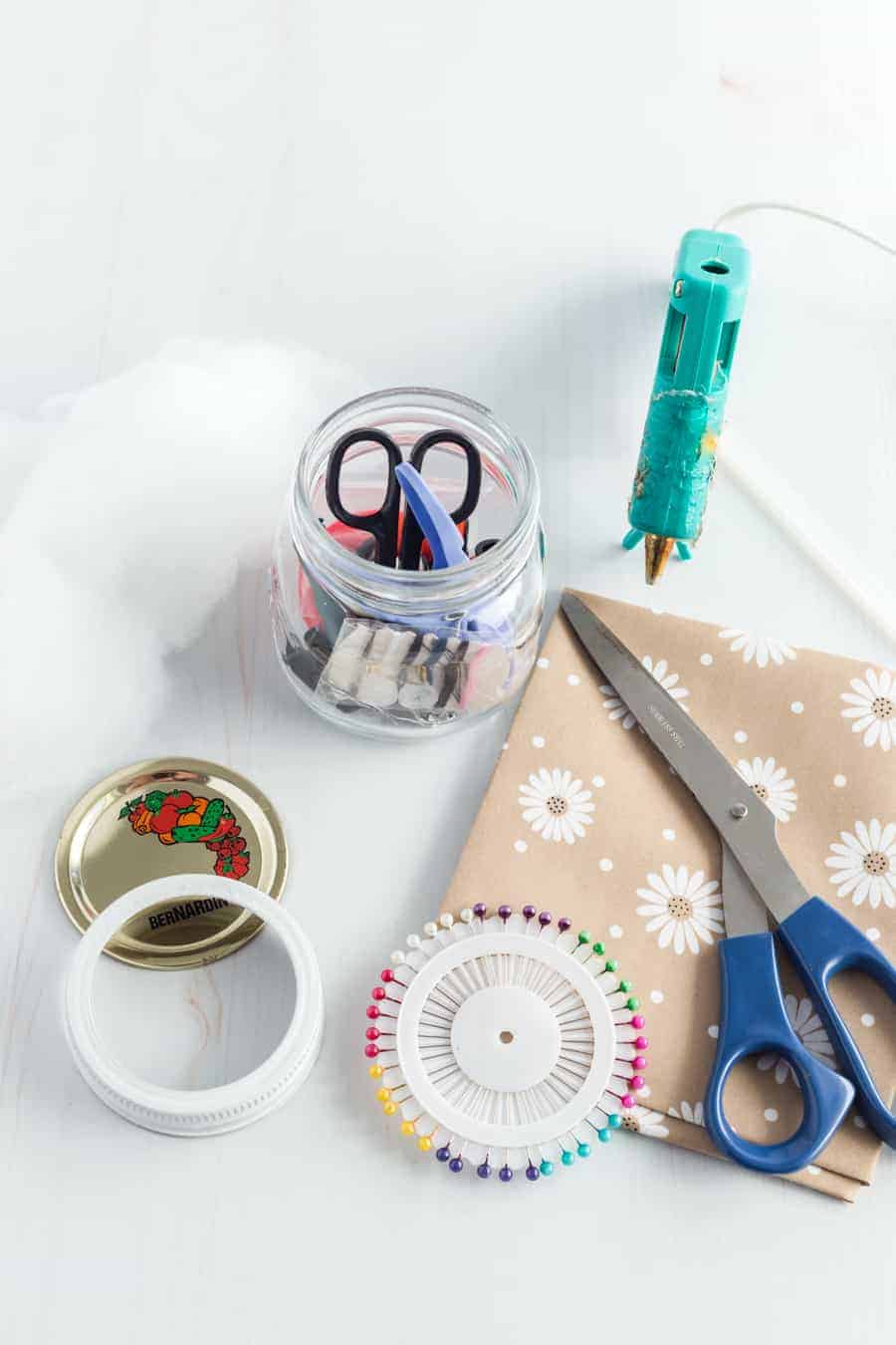 A clear jar with some sewing tools in it and the lid laid out. Some paper with scissors on it and glue gun nearby, as well as some pins.