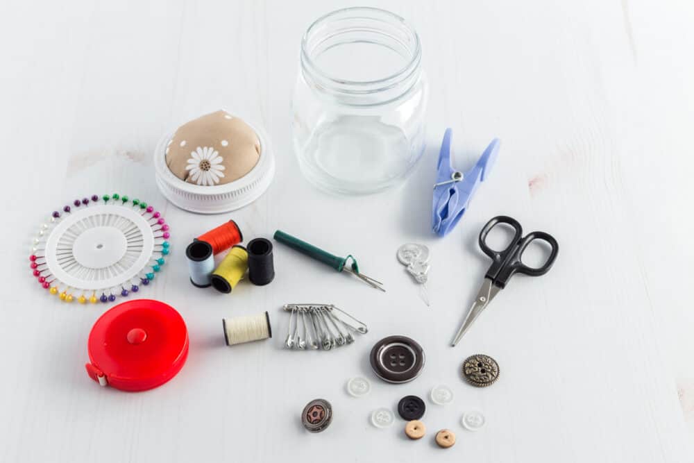 A small clear jar with sewing stuff around it, including, scissors, seam ripper, buttons, different color thread, a tape measure, and pins.