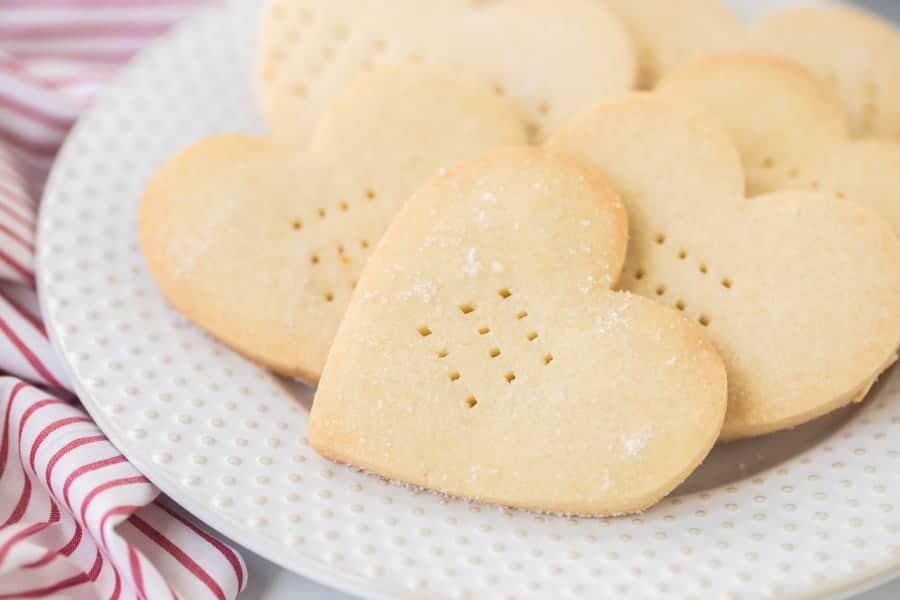 A close up of shortbread heart shaped cookies on a decorative white plate, on top of a red and white striped dish towel.