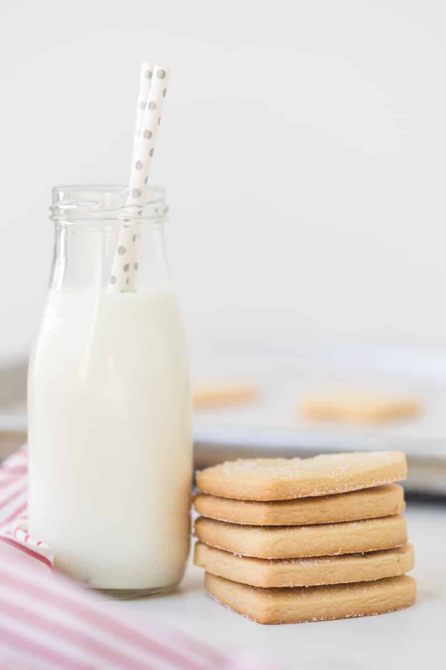 A glass jug of milk with two white straws polka dotted with silver, next to a stack of heart shaped shortbread cookies.