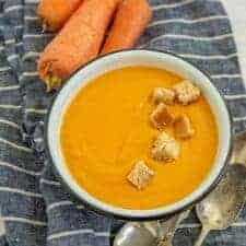 photo of a bowl of creamy carrot soup with a row of croutons in it a raw whole carrots in the background