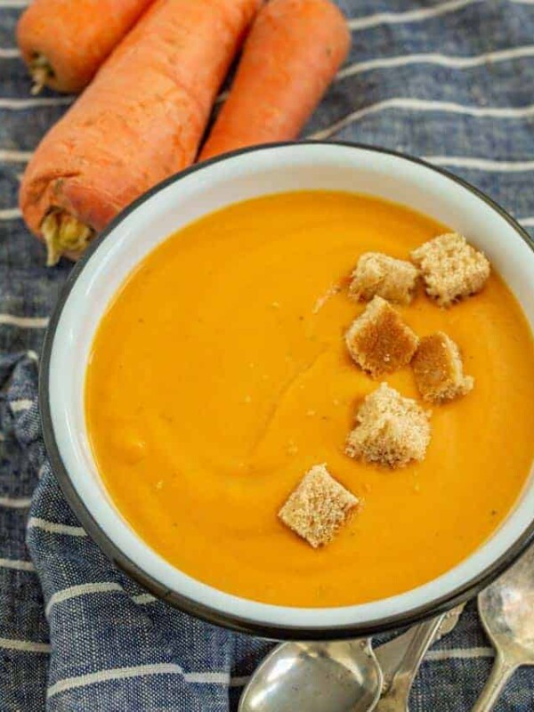 photo of a bowl of creamy carrot soup with a row of croutons in it a raw whole carrots in the background