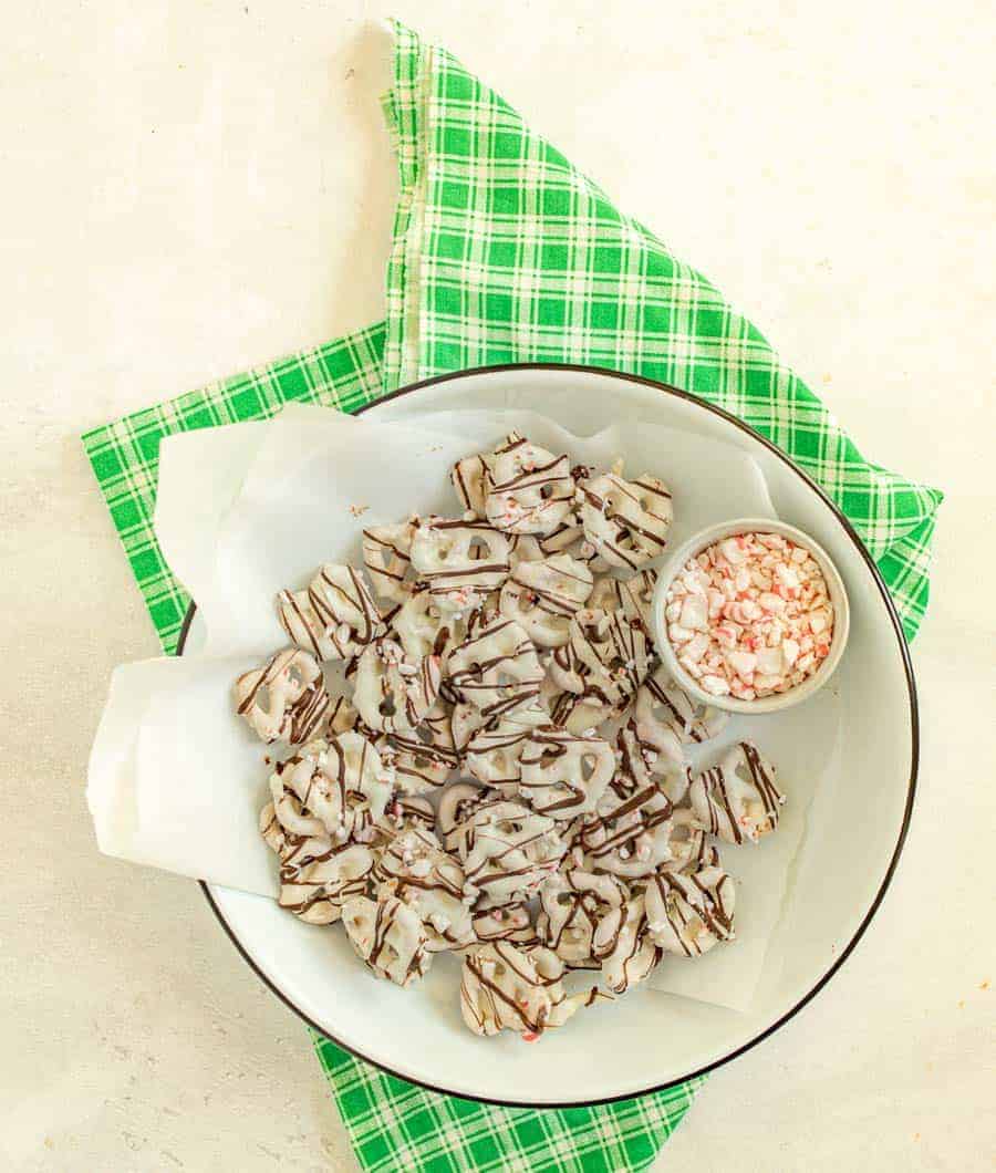 An overhead shot of white chocolate covered pretzels drizzled in chocolate by a bowl of crushed peppermint pieces on top of a plate, on top of a green and white checkered dish towel.