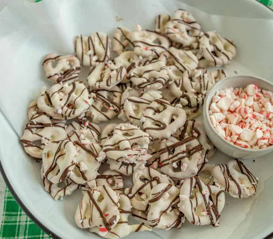 Close up of a plate of white chocolate covered pretzels striped with chocolate and small pieces of peppermint sprinkled on. There is also a small bowl of broken up peppermint pieces.
