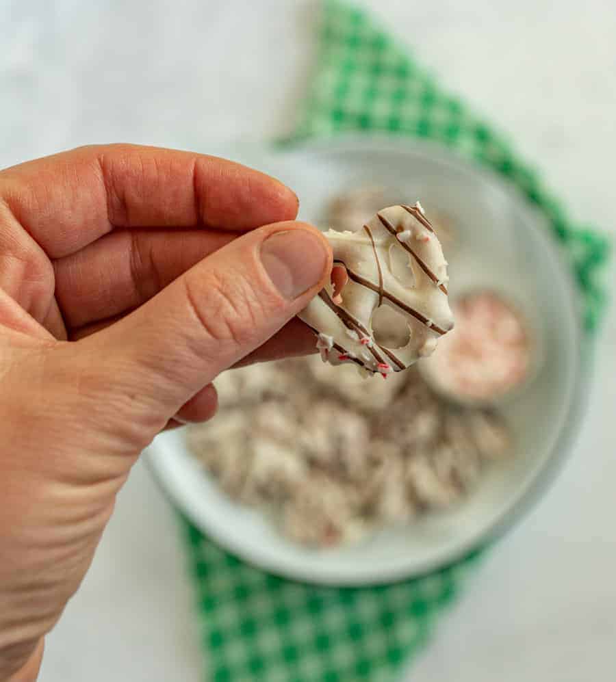 In the foreground is a person holding a pretzel that has been dipped in white chocolate, has chocolate stripes on it and sprinkles. In the background, there is a larger bowl full of the pretzels on top of a green and white checkered towel.