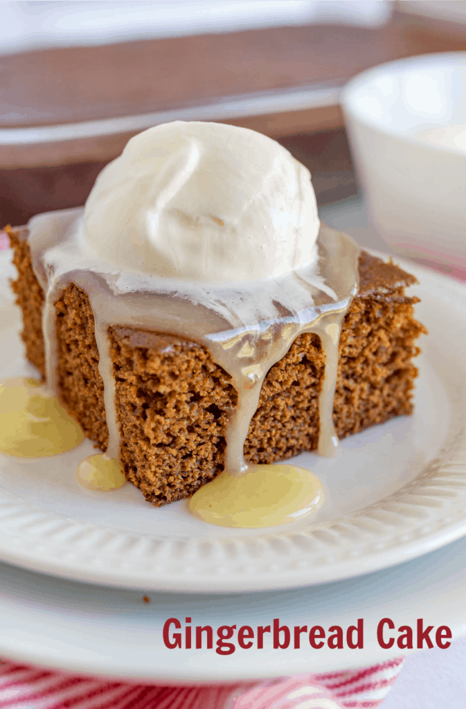 A square piece of gingerbread cake on a white plate topped with vanilla caramel sauce and whipped cream