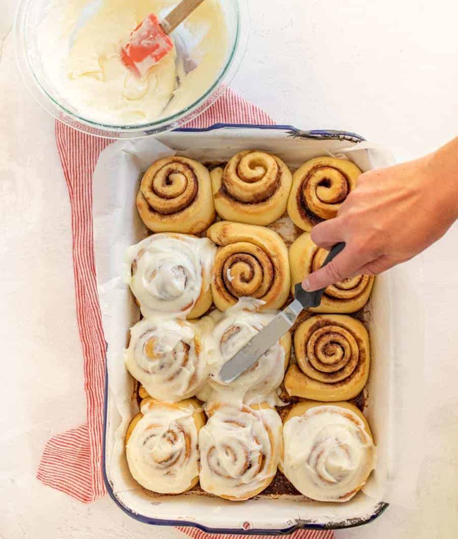 An overhead shot of an 11x13 pan full of cooked cinnamon rolls being frosted by someone. In the top left corner is a bowl full of icing and a spatula.