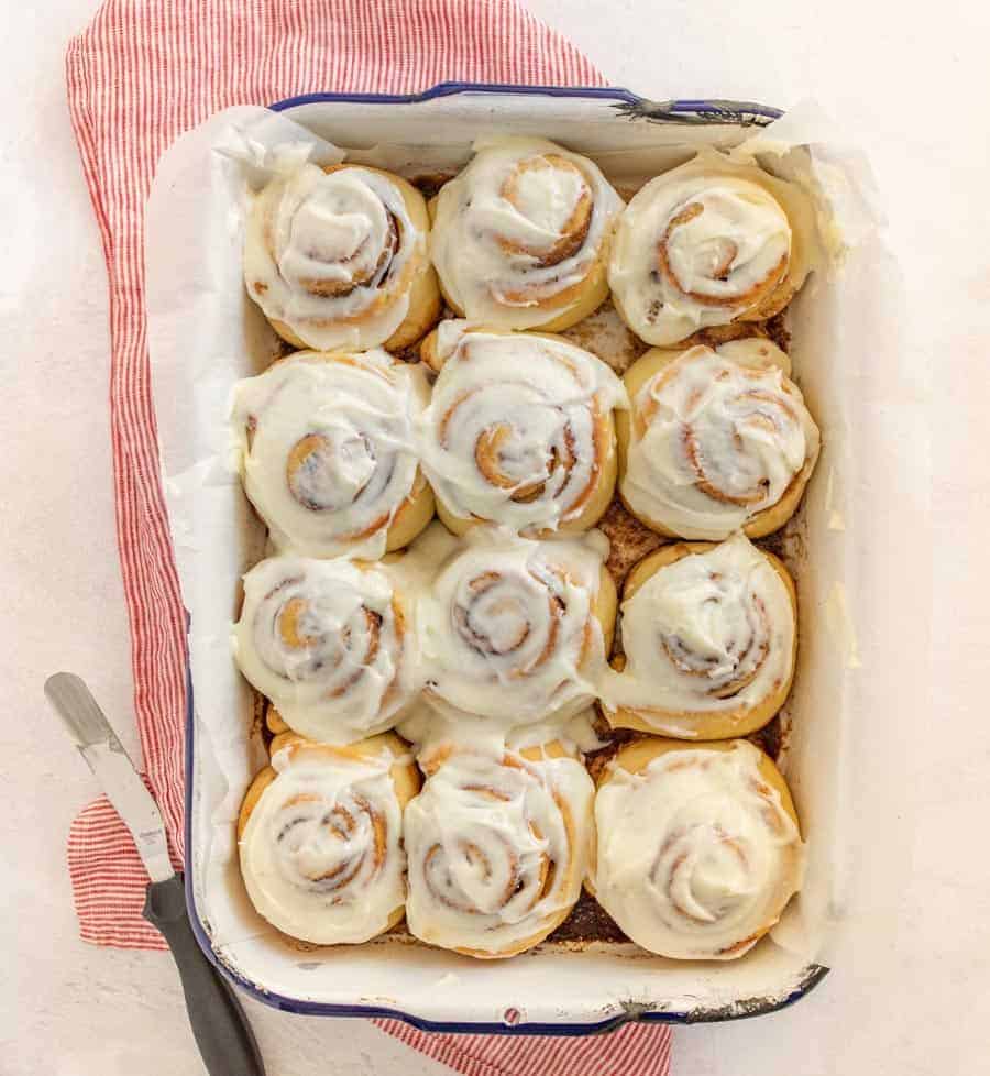 An overhead shot of the finished cinnamon rolls in a pan on top of a red and white striped towel. The bottom left-hand corner has the utensil used to frost the cinnamon rolls.