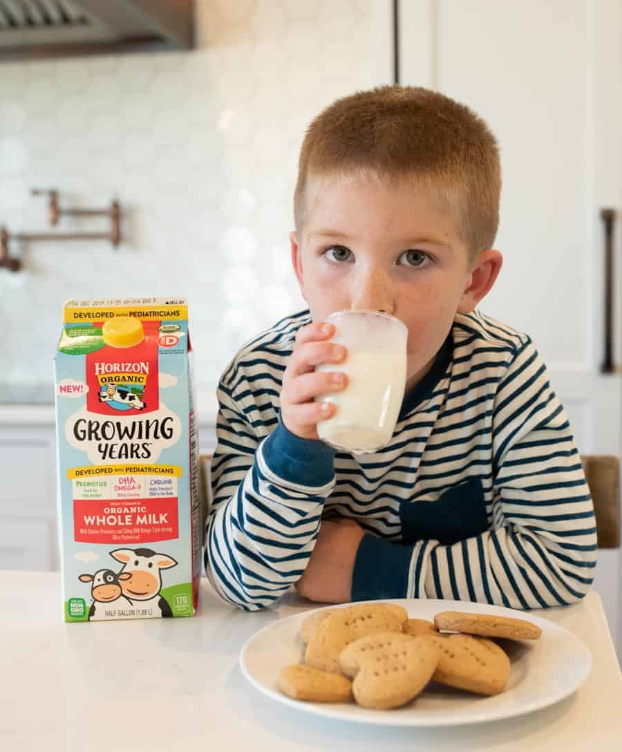 little boy with brown hair wearing a blue and white striped shirt drinking a glass of milk with a plate of heart-shaped graham crackers in front of him.