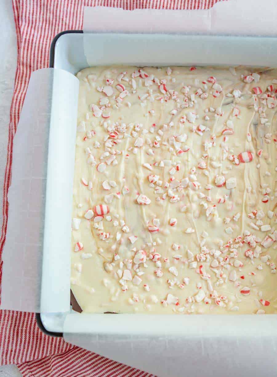 An up close shot of peppermint bark in a white square pan with a green outline, resting on top of a red and white striped towel.