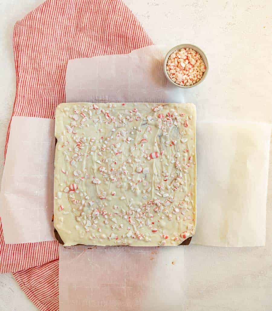 An overhead shot that in the middle has a large square piece of peppermint bark resting on a red and white striped towel. In the upper right hand side is a bowl full of peppermint pieces.