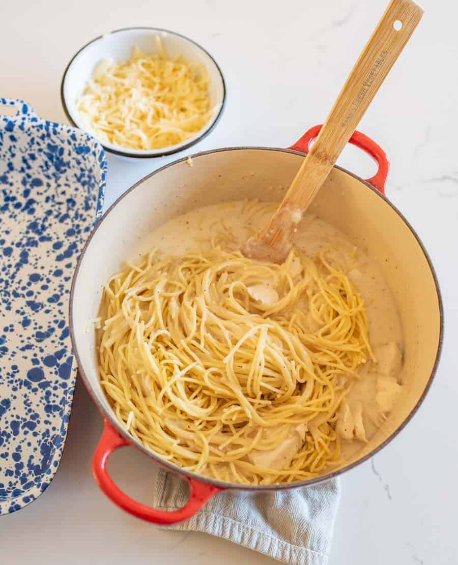 An overhead shot of the noodles and other ingredients being stirred with a wooden utensil in a ceramic bowl with two red handles. A bowl of cheese is in the upper left-hand corner waiting to be added to the recipe.