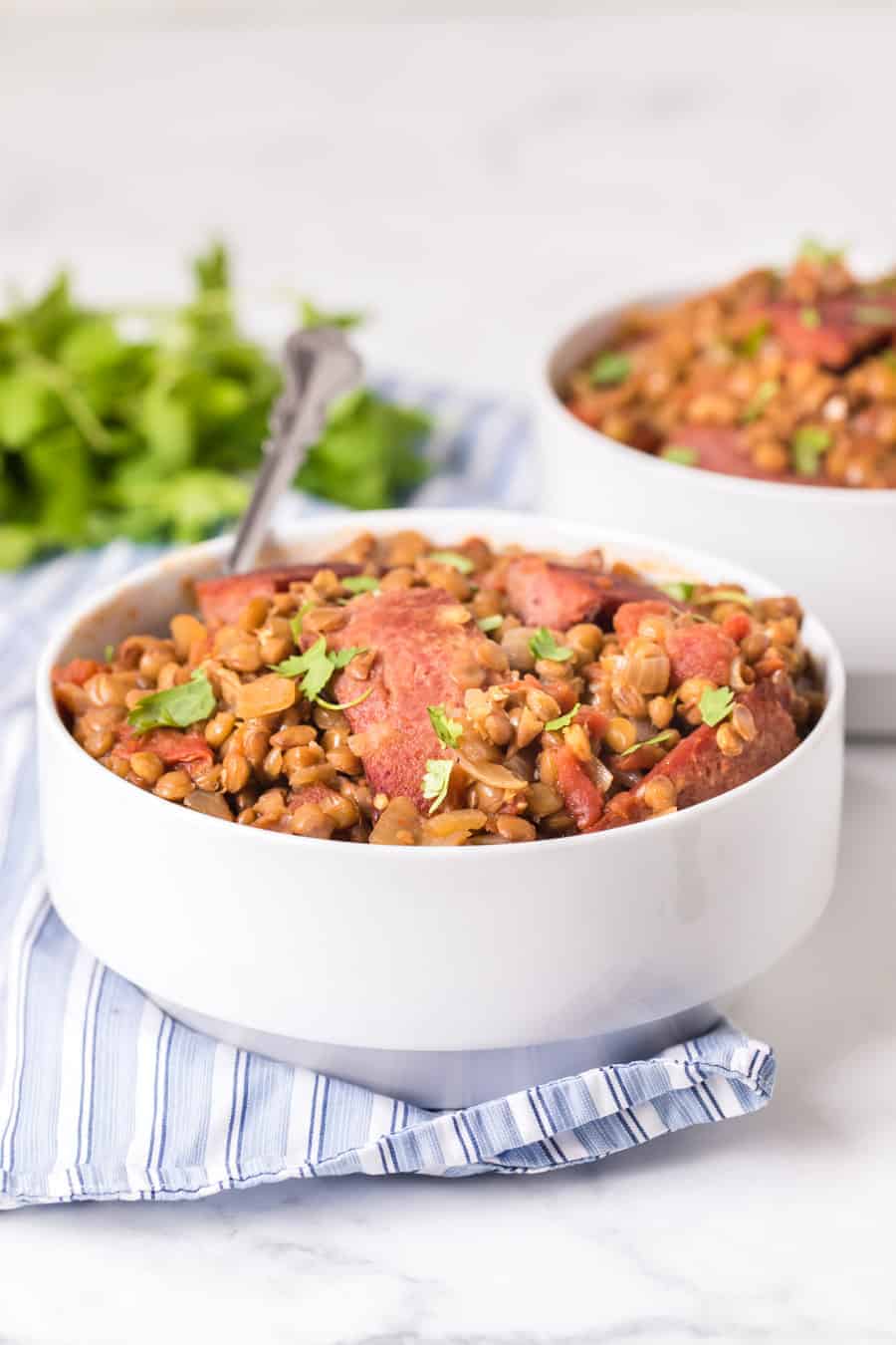 two white bowls of spicy lentils with sausage on blue striped towel