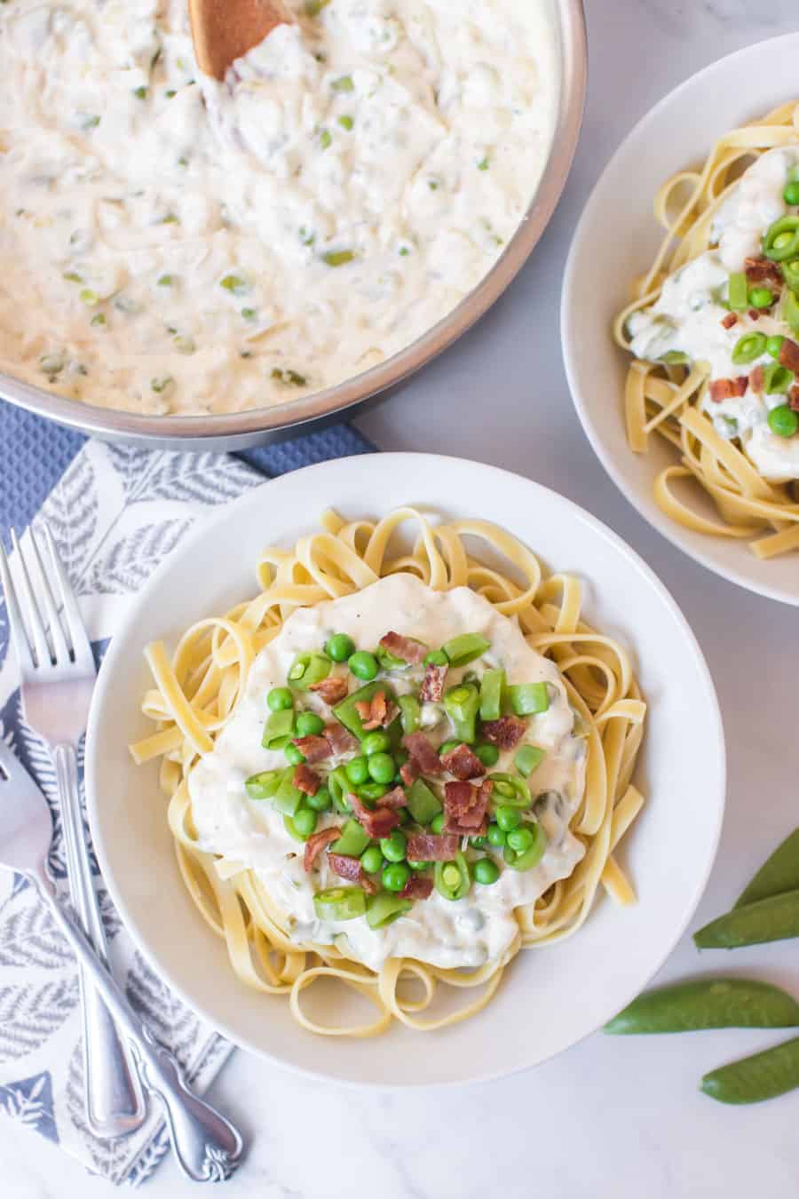 two round white bowls of creamy bacon and peas pasta next to pot of 